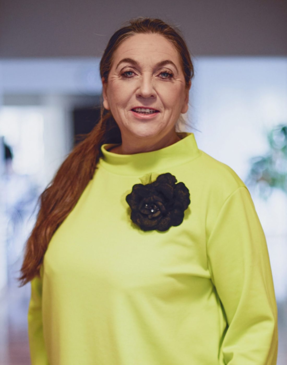 Anna_Jochim_Labuda Na zdjęciu znajduje się kobieta z długimi, rudymi włosami w bluzie z długim rękawem i kwiatem. Stoi przodem, widać ją od pasa w górę, patrzy na wprost.